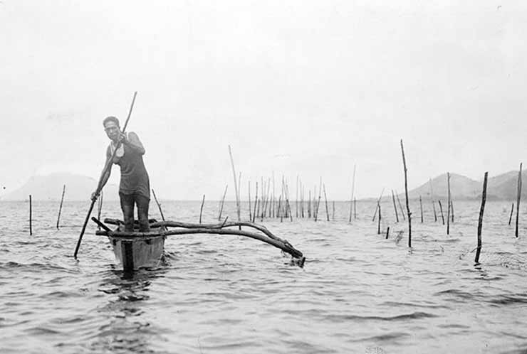 A man standing in an outrigger canoe, using a pole to propel it through the water